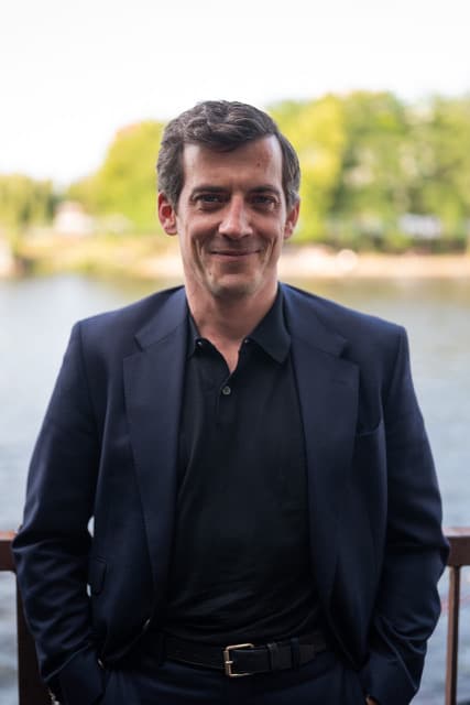 Nicolas Baum schaut lächelnd in die Kamera. Im Hintergrund sind ein Gewässer und Bäume erkennbar.