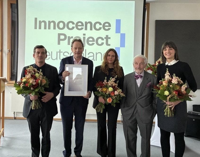 Bei der Verleihung des Peter-Raue-Preises 2025 stehen Tatjana Holter, Laura Farina Diederichs und Nicolas Baum jeweils einen Blumenstrauß haltend neben Stefan König, der die Siegerurkunde hält und Peter Raue. Alle blicken zur Kamera.