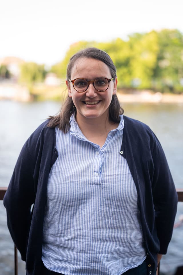 Carolin Arnemann schaut lächelnd in die Kamera. Im Hintergrund sind ein Gewässer und Bäume erkennbar.