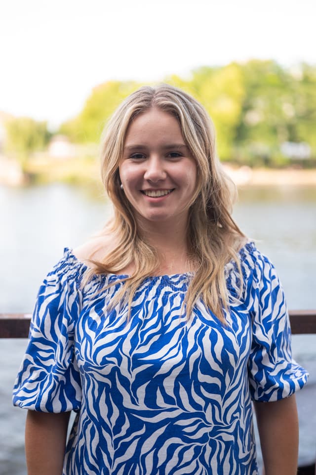 Clara Eckardt schaut lächelnd in die Kamera. Im Hintergrund sind ein Gewässer und Bäume erkennbar.