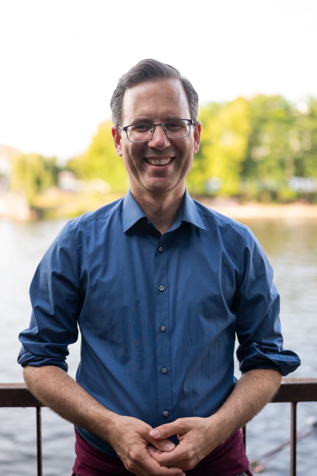 Johannes Kaspae schaut lächelnd in die Kamera. Im Hintergrund sind ein Gewässer und Bäume erkennbar.