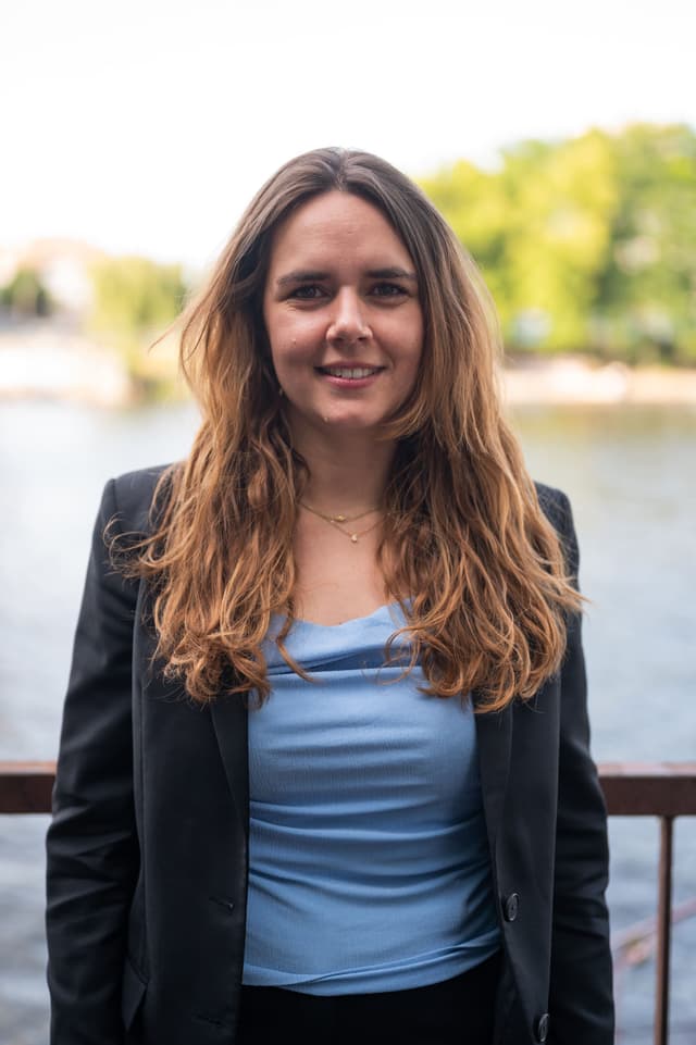 Laura Farina DIederichs schaut lächelnd in die Kamera. Im Hintergrund sind ein Gewässer und Bäume erkennbar.